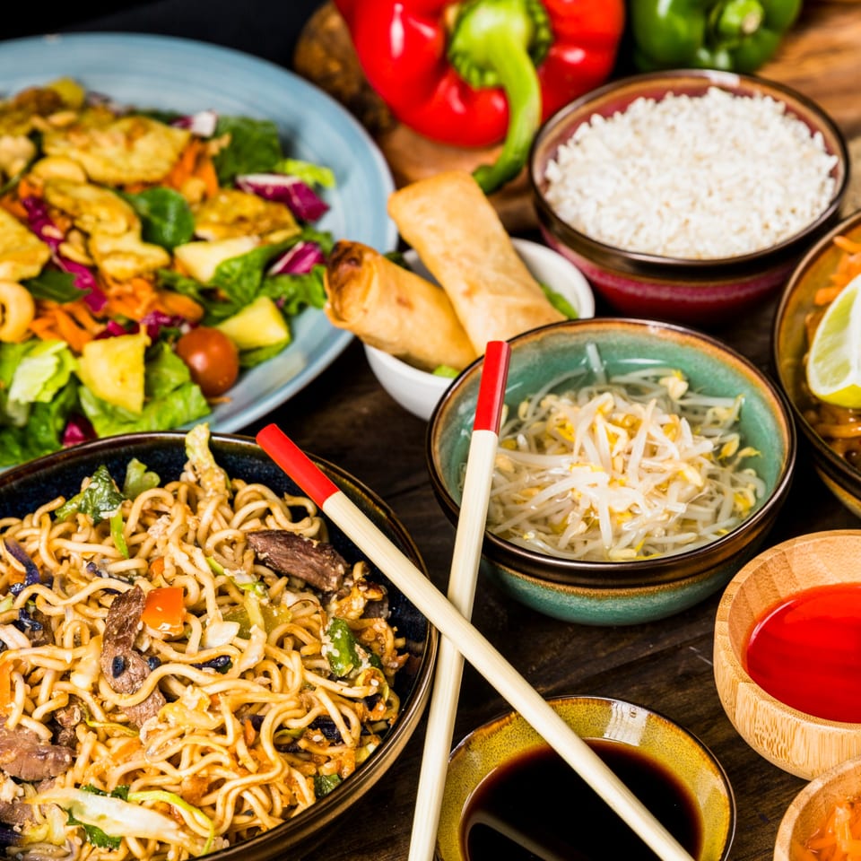 An overhead view of various Chinese dishes including noodles, rice, spring rolls, and salad.