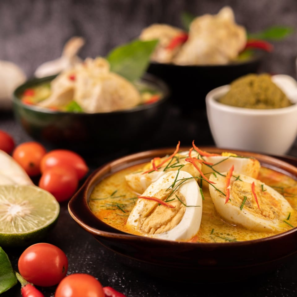 Curried hard-boiled eggs in a brown bowl, garnished with chili and herbs, with other dishes in background.