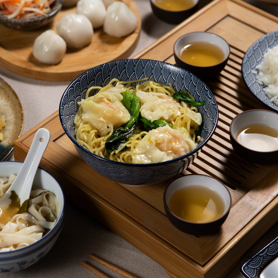 A bowl of wonton noodles with bok choy, tea, rice, and dumplings on a wooden tray.